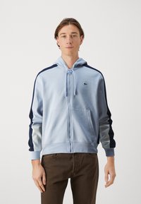 Svetlomodrá mikina na zips s tmavomodrými bočnými pruhmi, kapucňou na šnúrku a prednými vreckami. Obsahuje malé vyšívané logo na hrudi.