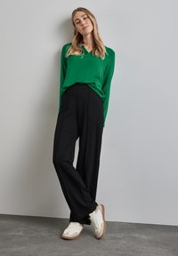 Maglietta verde a maniche lunghe con scollo a V abbinata a pantaloni larghi neri a vita alta e sneakers bianche con dettagli marroni su sfondo grigio.