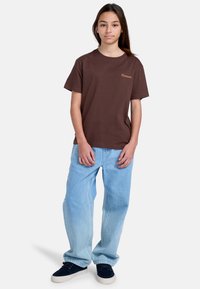 Brun bomullströja med korta ärmar och rundad halsringning, med en subtil guld "Element"-logotyp. Tillsammans med ljusblå baggyjeans.