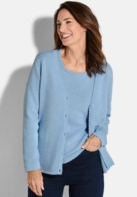 Hellblauer Cardigan aus strukturiertem Stoff, mit rundem Ausschnitt, Knopfverschluss und gewelltem Saum. Getragen über einem passenden Oberteil und dunklen Hosen.