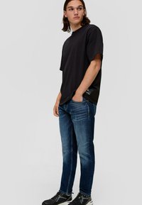 Schwarzes Baumwoll-T-Shirt mit lässigem Schnitt, kombiniert mit dunkelblauen Jeans. Das Model trägt schwarze Sportschuhe und steht vor einem neutralen Hintergrund.