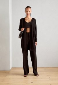 Un long cardigan noir à la coupe décontractée, porté par-dessus un haut court et un pantalon large. Les accessoires comprennent un petit sac à épaule et des bijoux.