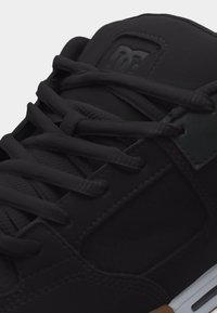 Close-up van een zwarte sneaker met zwarte veters, gevoerde tong met het DC-logo in reliëf en een witte tussenzool met bruine gumzool.