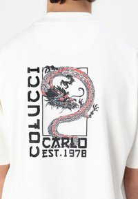 Hvit t-skjorte med et stort, fargerikt dragemotiv på baksiden, med fet svart tekst som leser "COLUCCI CARLO EST. 1978."