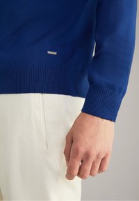 Gros plan sur une personne portant un pull bleu à manches longues avec l'étiquette "JOOP!" et un pantalon blanc cassé, la main détendue le long du corps.