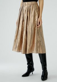 Metallic bronzen midi-rok met plooien, met een gathered tailleband. Gecombineerd met kniehoge zwarte nauwsluitende laarzen. Gladde, reflecterende textuur.
