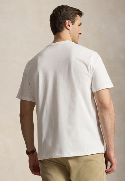 Mann trägt ein schlichtes weißes Kurzarm-T-Shirt und beige Hose, steht mit dem Rücken zur Kamera vor neutralem Hintergrund.