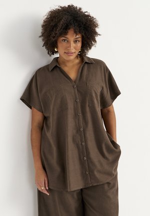 Kvinde med krøllet hår iført løs brun skjorte med knapper og matchende bukser, står mod hvid baggrund med hånden i lommen.