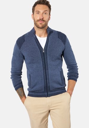 Mann mit Bart, der einen blauen Reißverschluss-Pullover über einem weißen Hemd und beigen Hosen trägt, Hände in den Taschen, stehend vor einfarbigem Hintergrund.