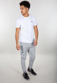Weißes Baumwoll-T-Shirt mit kleinem schwarzem Logo, kombiniert mit grauen Jogginghosen mit schwarzem Markennamen-Druck und schwarzen Turnschuhen.