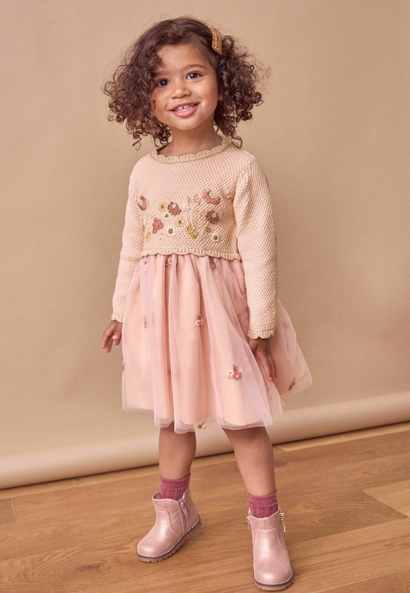 Petite fille souriante aux cheveux bouclés porte une robe fleurie rose, un pull en tricot, des chaussettes roses et des bottines brillantes sur un sol en bois.