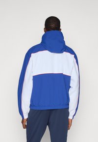 Blå og hvit hettejakke med fargiblokkmønster, med elastiske mansjetter og linning, og tynne røde detaljer på sømmene.