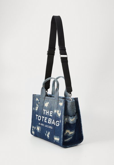Borsa tote in denim con Toppa usurata, tracolla nera regolabile e doppi manici superiori. Presenta testo bianco: "THE TOTE BAG M A RC JACOBS."