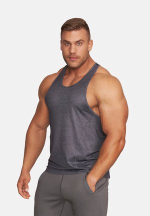 Muskelbepackter Mann trägt ein dunkelgraues Tanktop und graue Hose, steht mit einer Hand in der Tasche vor einem einfarbigen Hintergrund.