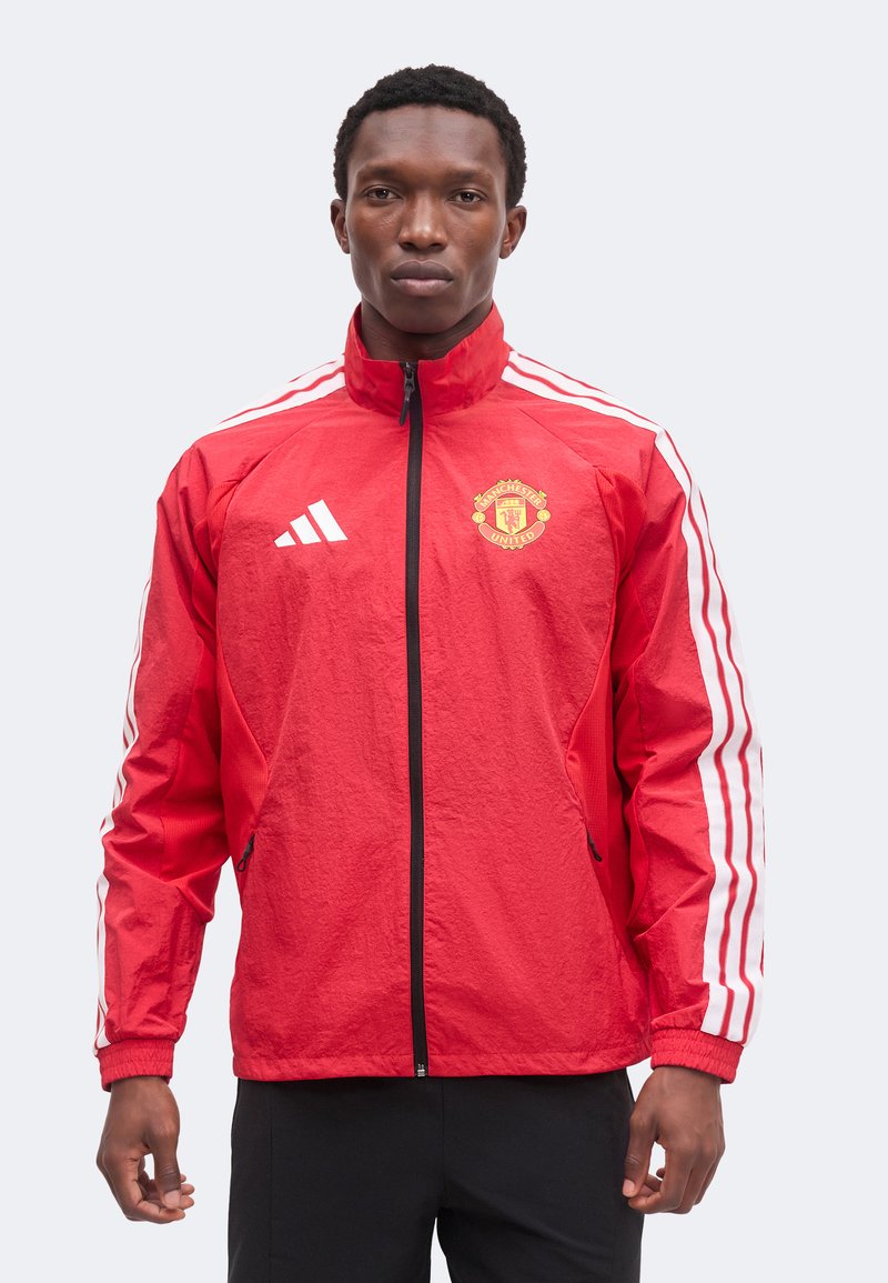 Άνδρας που φορά κόκκινο μπουφάν Adidas της Manchester United με λευκές ρίγες στα μανίκια, στέκεται μπροστά σε ανοιχτό φόντο.