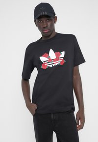 Fekete rövid ujjú póló, amelyen az elején fehér Adidas logó található, piros rózsákkal és zöld levelekkel díszítve. Kényelmes szabású, kerek nyakkivágással.