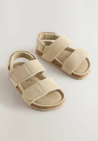 Beige Wildledersandalen mit zwei verstellbaren Klettverschlüssen, offener Rückseite und strukturierten Gummisohlen. Flaches Design mit minimalistischem Ästhetik.