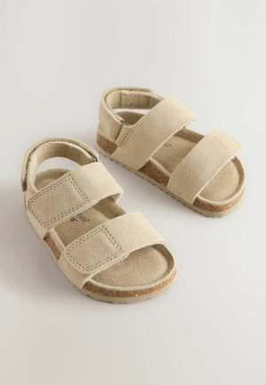 Beiga mockaskor med två justerbara kardborreband, öppen baksida och texturerade gummisulor. Platt design med en minimalistisk estetik.