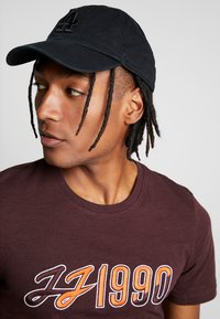 Gorra negra con logo bordado, combinada con una camiseta texturizada color burdeos con diseño gráfico en naranja y blanco, de cuello redondo.