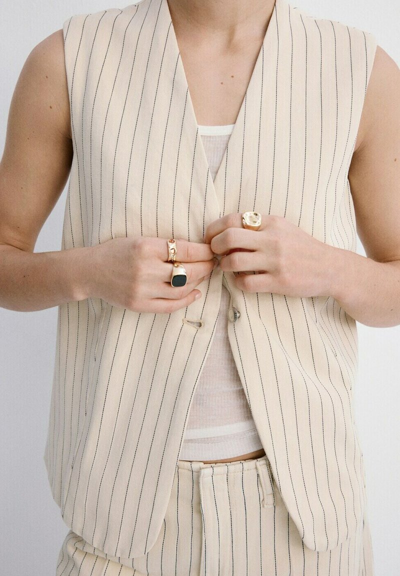 Personne portant un gilet beige à fines rayures et un pantalon assorti, en train de boutonner le gilet, avec trois bagues en or visibles aux doigts.