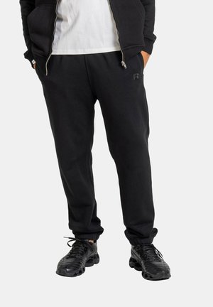 Man wearing black sweatpants, black sneakers, white T-shirt, and black zip-up jacket with hands in pockets against white background.