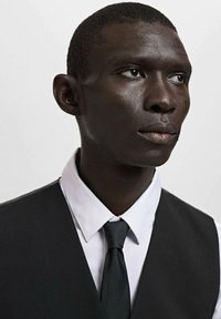 Schwarzer Smoking mit weißem Hemd, ausgestattet mit einer schwarzen Weste und Krawatte. Glatter Stoff, figurbetontes Design und dezente Texturdetails.