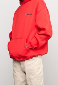 Persona che indossa una felpa rossa vivace con la scritta "MULTIPLY" sul petto, mani nella tasca anteriore, abbinata a pantaloni beige chiaro.