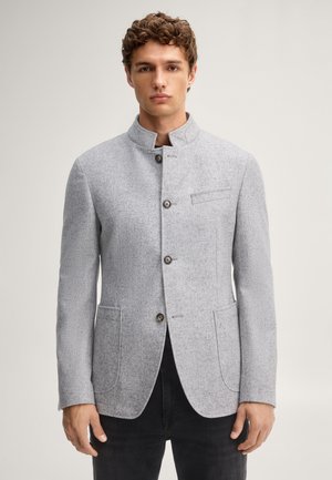 Junger Mann trägt eine hellgraue, geknöpfte Wolljacke mit Stehkragen und schwarze Hose, schaut nach vorne vor einem einfarbigen Hintergrund.