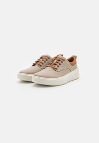 Zapatillas de lona beige con detalles en cuero marrón, suela de goma blanca y cordones a juego. Presentan una superficie texturizada y un diseño de punta redondeada.