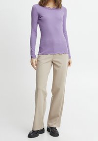 Blusa de manga larga de color púrpura claro con un escote y bordes de mangas en forma de ondulación, combinada con pantalones de pierna ancha beige y zapatos chunky negros.