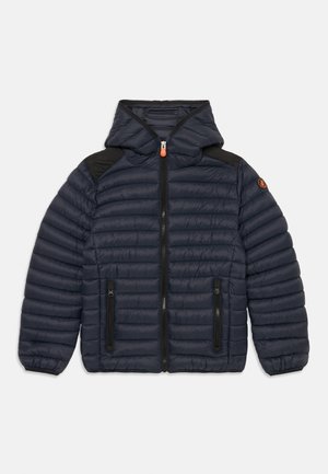 Chaqueta acolchada azul marino con capucha, que presenta tejido acolchado, acentos negros en los hombros y bolsillos con cremallera en la parte delantera. Detalle de tirador de cremallera en color naranja.