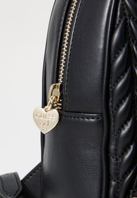 Sac à dos en cuir noir avec texture matelassée, fermeture éclair dorée et un charm en forme de cœur portant l'inscription "GUESS Girl" avec un petit accent en pierre précieuse.