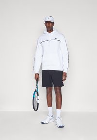 Hanorac alb cu glugă și dungi negre, pantaloni scurți negri, adidași albi și o șapcă albă cu un logo verde, ținând o rachetă de tenis.