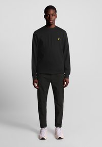 Giovane in piedi che indossa una maglietta nera a maniche lunghe con un logo giallo, pantaloni neri e sneaker bianche su uno sfondo grigio semplice.