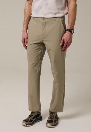 Hombre vestido con pantalones casuales beige, reloj de pulsera negro y zapatos deportivos grises y negros, de pie sobre un fondo neutro.