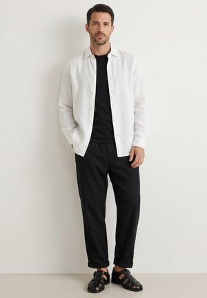 Mand stående mod en hvid væg iført en hvid knapforan skjorte over en sort T-shirt, sorte bukser og sorte lædersandaler.