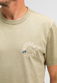 Lichtbeige katoenen T-shirt met ronde hals. Heeft een cirkelvormige logo-print in het wit en blauw met de tekst "BUTCHER OF BLUE" op de borst.