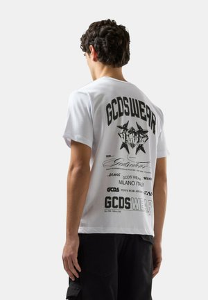 Modello maschile di spalle che indossa una maglietta bianca GCDSWEAR con molteplici scritte grafiche nere e design sul retro, abbinata a pantaloncini neri.