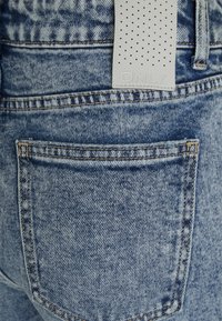 Jeans de denim azul claro com um acabamento texturizado. Apresenta um bolso traseiro, costura visível e uma etiqueta de couro cinza com marca.