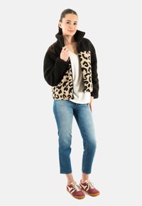 Giacca di pile con motivo leopardato nero e marrone chiaro, colletto alto, chiusura a zip; indossata con jeans chiari e sneakers rosse. Texture morbida.