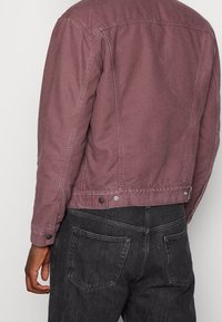 Blek mauve denimjacka med knappdetaljer, synlig sömnad och en skräddarsydd passform; bärs över mörka jeans i en avslappnad stil.