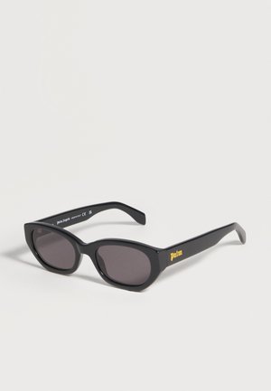 Lunettes de soleil noires forme œil de chat avec verres foncés et « palm » écrit en jaune sur la branche, présentées sur un fond clair uni.