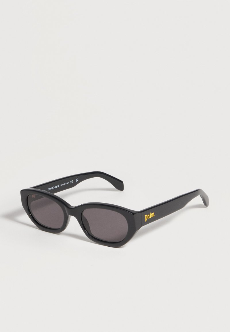 Lunettes de soleil noires forme œil de chat avec verres foncés et « palm » écrit en jaune sur la branche, présentées sur un fond clair uni.