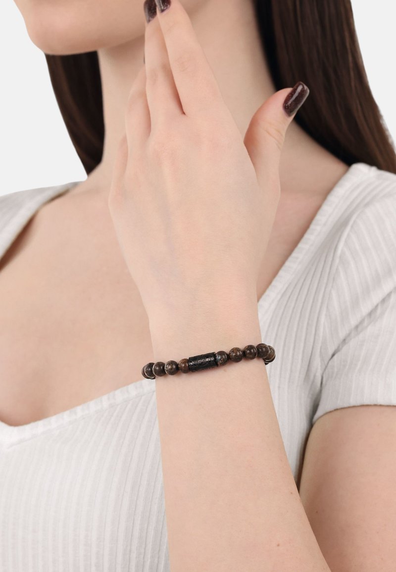 Bracciale di perline con pietre rotonde marroni e una perla nera centrale, indossato su un polso con uno sfondo di camicia chiara.