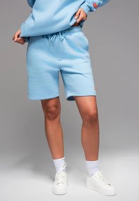 Shorts en coton bleu clair avec taille élastique et cordon de serrage. Dispose de poches latérales et d'un petit logo sur l'ourlet. Portés avec des chaussures et des chaussettes blanches.