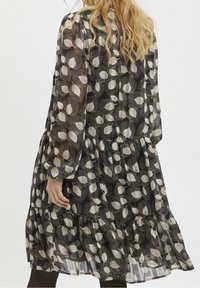 Robe noire à manches longues, ornée d'un motif de feuilles en crème et beige. La jupe présente un design à volants superposés et une texture en tissu transparent.