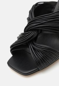 Sandale à talon noir avec des brides en caoutchouc torsadées, un bout carré et une semelle texturée. Le design met en avant une esthétique moderne et minimaliste.