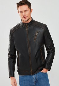 Chaqueta de cuero negro con cuello alto, acentos acolchados en los hombros, bolsillos con cremallera y detalles de costura marrón. Acabado texturizado.
