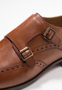 Chaussure monk en cuir marron avec deux boucles en laiton et perforations décoratives, gros plan sur le dessus et les détails latéraux.