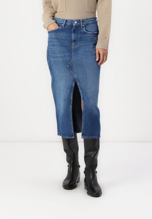 Personne portant un pull beige, une jupe midi en jean bleu avec une fente à l'avant, et des bottes noires hautes jusqu'aux genoux, debout devant un fond uni.